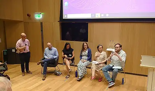 Five people seated, one speaking into a microphone. A sixth person is stood to the side holding another microphone.