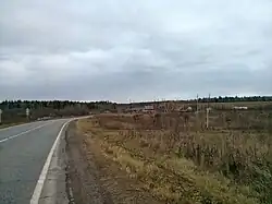 A paved road on the left with a field on the right. The field is filled with green and brown grass and the sky is overcast.