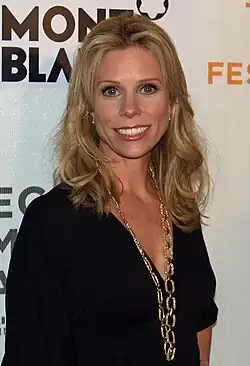 A blonde Caucasian woman wearing a black dress and long gold necklace