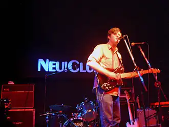 blurry image of Chris Bathgate wearing a short-sleeved checkered shirt and a pair of jeans, singing and playing electric guitar on a dimly lit stage, with a drum kit and speakers in background and a neon sign reading "NEU!CLUB"