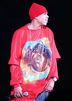 A man in a red hat, wearing a long red t-shirt over a red long-sleeved shirt, holding a microphone in his right hand.