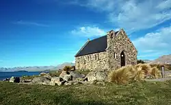 A small stone church by a lake surrounded by a large grassy area with some boulders