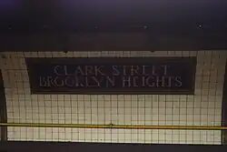 A dark tile mosaic on the curved wall of the station, reading "Clark Street Brooklyn Heights". The rest of the wall is clad with white tile.