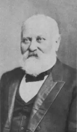 portrait shot of a man in late middle age with white hair and beard. He is seen to wear a suit with vest and a small bow-style tie.