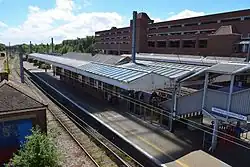 A view of a railway island platform with overhead wires above the tracks from the footbridge to the left hand side. On the side of the stairs to the platform down the footbridge is a sign that reads "Welwyn Garden City", and there is also a brick platform building with a large canopy extending over the platform.