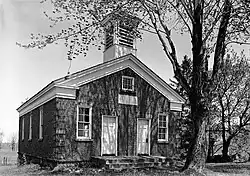 A black and white image of a building surfaced in small round stones, seen from slightly to its left. It has a pointed roof with a square cupola on top in the front. A large tree is in front of it, partially obscuring the view