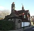 Almost side-on view of a long, two-storey house with two gable ends, large areas of tiles, a white ground floor and a flint section nearest the camera (with arched recess and brick buttress). Two tall chimneys rise from the roof. A low, narrow pavement with bollards runs alongside.