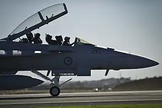 Forward section of military jet on an airfield, with open canopy and two crewmen seated in the cockpit
