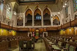 The House of Commons, divided into opposing ranks of desks, where the government faces the opposition across the floor; the Speaker's chair is at head of the aisle, between the two ranks of desks; green decor