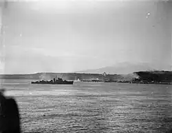 A black and white photograph showing a warship firing its armament at positions on the foreshore, while in the distance landing craft move towards the beach