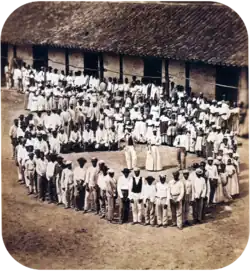 Photograph showing a group of people dressed in white, who have gathered in front of a tile-roofed farm building and observe another large group which has formed a large circle surrounding 5 men straddling large drums, a woman and 2 other men.