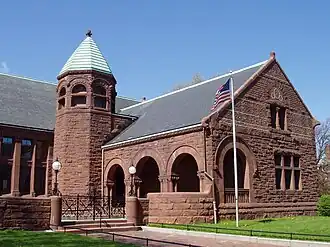 A brownstone building with arched porch. The roof is slate, except for a tower, which has a green copper roof. A gate in front of the building is made of ornate wrought iron, which is also present in globular light fixtures mounted on either side.