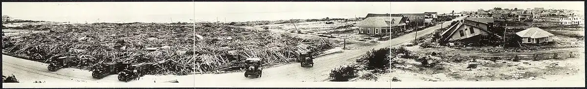 Panorama showing wooden buildings reduced to heaps of wooden planks along the waterfront. Other buildings remain standing and show varying degrees of damage.