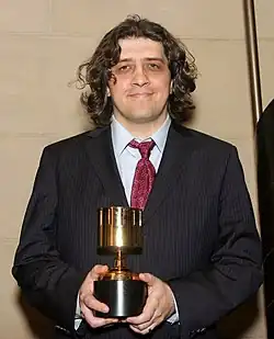 A man with short, curly brown hair wearing a black suit with a red tie holding an Annie Award, facing forward.