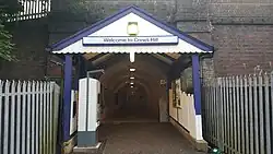 A blue portico archway is the entrance to a brick underpass with a brick wall on either side. There are metal fences on either side of the path. Above the archway a sign reads "Welcome to Crews Hill"