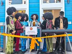 The Curtis Family C-Notes, a group with two adults and their five teenaged children, perform outdoors. The children all sport large afro hairstyles. Maestro Curtis plays the keyboard while Nola and their children sing, standing in a semicircle behind a microphone and a podium with the name and logo of the Community Music Center. A large orange ribbon is tied in front of the podium.