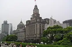 The Bund area of Shanghai. Pedestrians walk before a row of trees and a series of tall buildings. A blue sky overhead is obscured slightly by several clouds.
