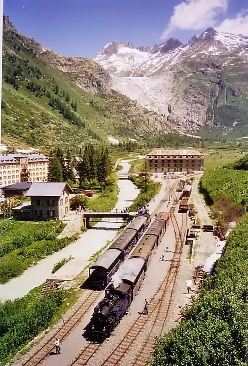 Two steam locomotive-pulled trains in a railway yard nestled in a valley