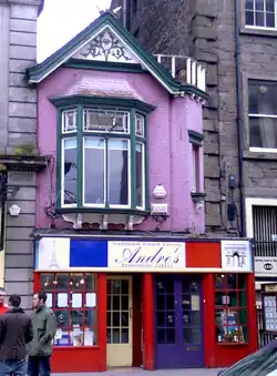 A two-floor building nestled between two larger houses. The lower half, housing a French restaurant, is painted brightly in blue, white, and red. The nondescript upper half has a brick facade in a faded pink colour and dark green wooden trims on the bay window and gable roof.