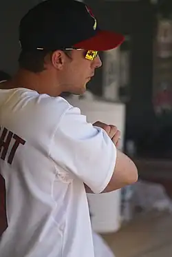 A man in a white baseball jersey, navy blue baseball cap with a red brim, and orange mirrored sunglasses stands with his arms crossed.