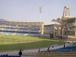 Back view of the entrance and a stand of a cricket stadium. Some people can be seen near the seats.