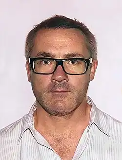 Damien Hirst, a man wearing glasses with brown rims, and a white shirt with faint vertical stripes