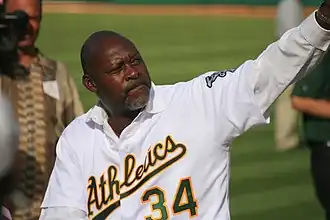 A man, now retired, in a white Oakland Athletics uniform, pictured close range from the waist up. His head is turned to his left, left arm waving, as if to a crowd in the stands.