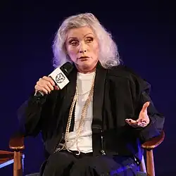 Debbie Harry, holding a microphone in her right hand and speaking into it; her left hand is outstretched in gesture