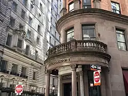 The building's entrance portico, which contains a curved balustrade supported by dark brown columns. There are windows above and next to the portico. A white building is in the background.