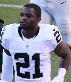 An American football player looks to his right. He is not wearing his helmet. He is wearing a white jersey with the number 21 across his chest in black.