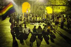 A yellow hued photo of people sitting a circle outside in plaza. A rainbow flag is waving off to the left