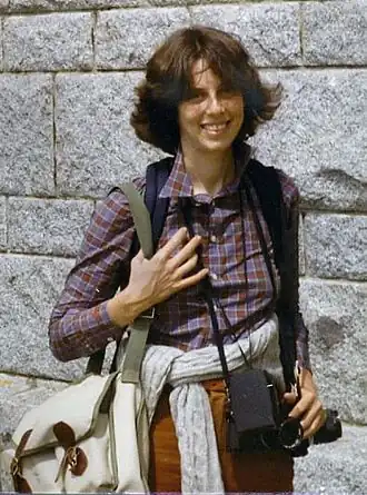 A photo of Diana Mara Henry standing with camera equipment draped over her shoulders.