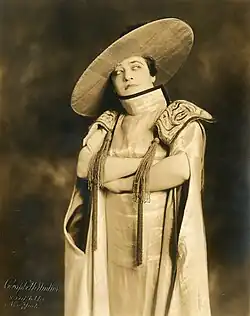 A white woman posing with her arms crossed, in a costume that includes a wide-brimmed headpiece, a high collar, and large shoulder pads