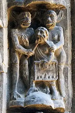 Sculpture of two horned grinning figures applying instruments to the face and tongue of a man holding a game board.