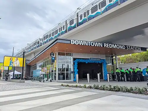 A train station entrance with a large sign reading "Downtown Redmond Station" mounted on a canopy. The entrance has large glass windows and is seen next to several scooters and bicycles.