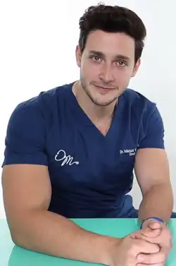 Headshot of Mike Varshavski. He is a strong, white man with dark brown hair, a stubble beard and moustache, and green eyes. He is wearing a T-shirt resembling medical scrubs with his name and logo, consisting of a stethoscope in the shape of the letter M.