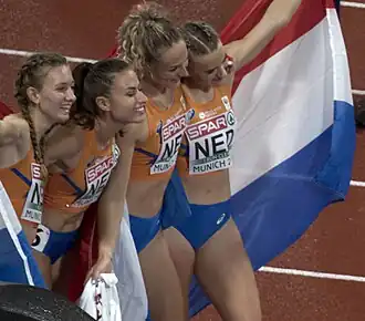 Photo of four women wearing orange tops and blue shorts holding red-white-and-blue flags