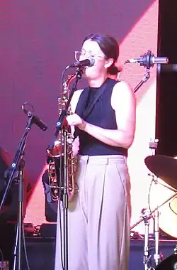 Jasmine Myra, wearing a black sleeveless top and grey pants, plays the alto saxophone on a stage behind a microphone