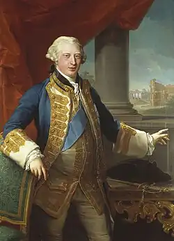 A three-quarter portrait of a young man. His hair is light grey/blonde. He wears pale leggings, a pale waistcoat decorated with gold lace, a large blue sash, and a blue and gold lace blazer. His right arm rests on a chair, his left hand points to a painting behind him.