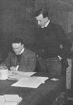 two middle-aged white men, both clean-shaven with full heads of neatly cut dark hair, the one on the left seated, the other standing