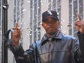 A dark-skinned man in a black baseball cap and black leather jacket holding up both his hands with his index fingers extended. He is holding a cigar in his right hand.