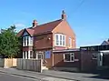 A converted early 20th-century house with bay windows and some black-and-white panelling. A low, flat-roofed extension on the near side has an entrance door above three concrete steps. A wooden fence obscures the lower half of the ground floor.