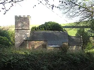 Stone building with tiled roof and square tower, surrounded by vegetation.