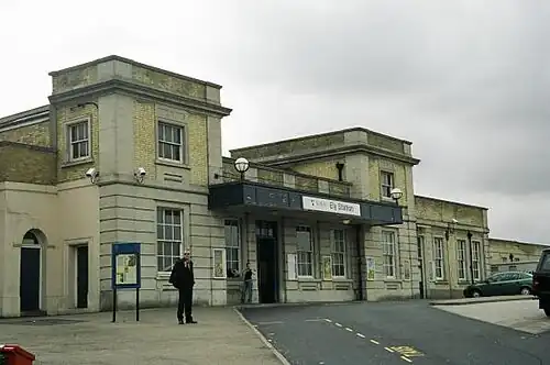 A white stucco building with a mix of one and two stories with sash windows as seen from the pavement a short distance away. Above the entrance is a sign that reads "Welcome to Ely" and a man is standing in front of the station.