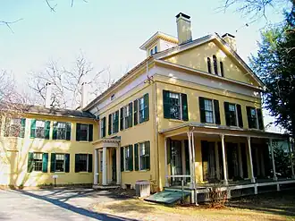 A yellow Federal style house with white trim and green shutters. The front of the house has a covered porch, but the main entrance is on the side of the house. It is protected by a small portico.