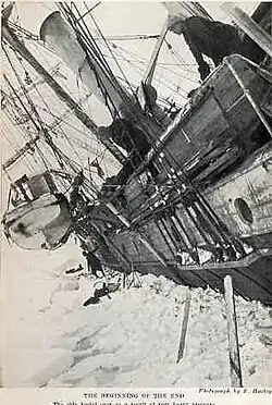 Side of a wooden steamship held in solid ice, leaning steeply to the left with a lifeboat swinging in its davits. One man visible on the ice, another aboard the ship, looking down.