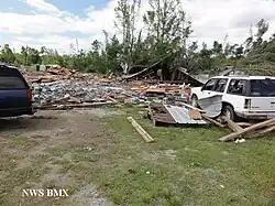 A building completely destroyed with trees in the background. Two cars are visible, one of which looks slightly damaged. On the bottom left, it says "NWS BMX".