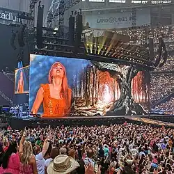A large screen on the Eras Tour displaying Taylor Swift, surrounded by her fans in the stadium