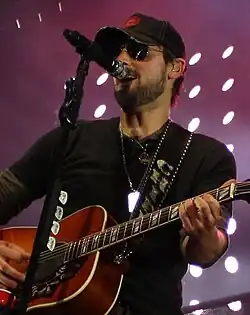 A man wearing a baseball cap, sunglasses and dark clothing, playing a guitar and singing into a microphone