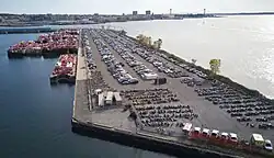 An aerial view of a L-shaped pier forming a harbor. The top of the pier has rows of parked automobiles. Several barges are moored.